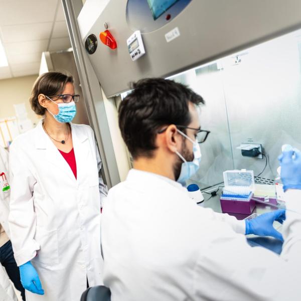 Two scientists standing in a wet lab watching a third, seated, pipetting
