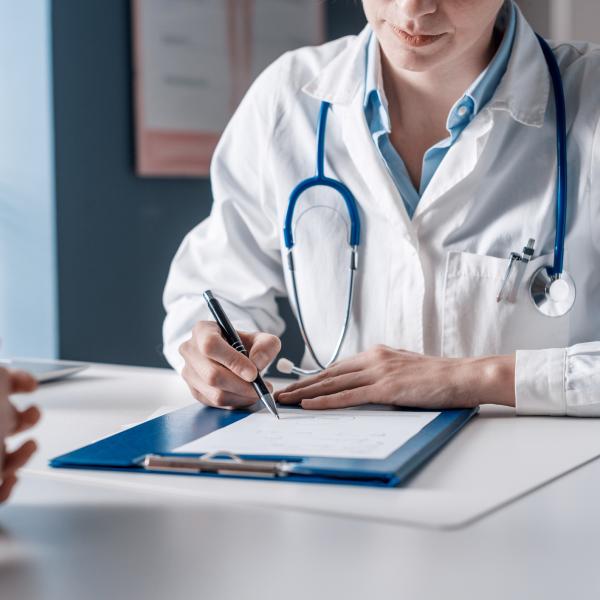 Doctor in white coat sitting at a desk making notes with a patient sitting across the desk